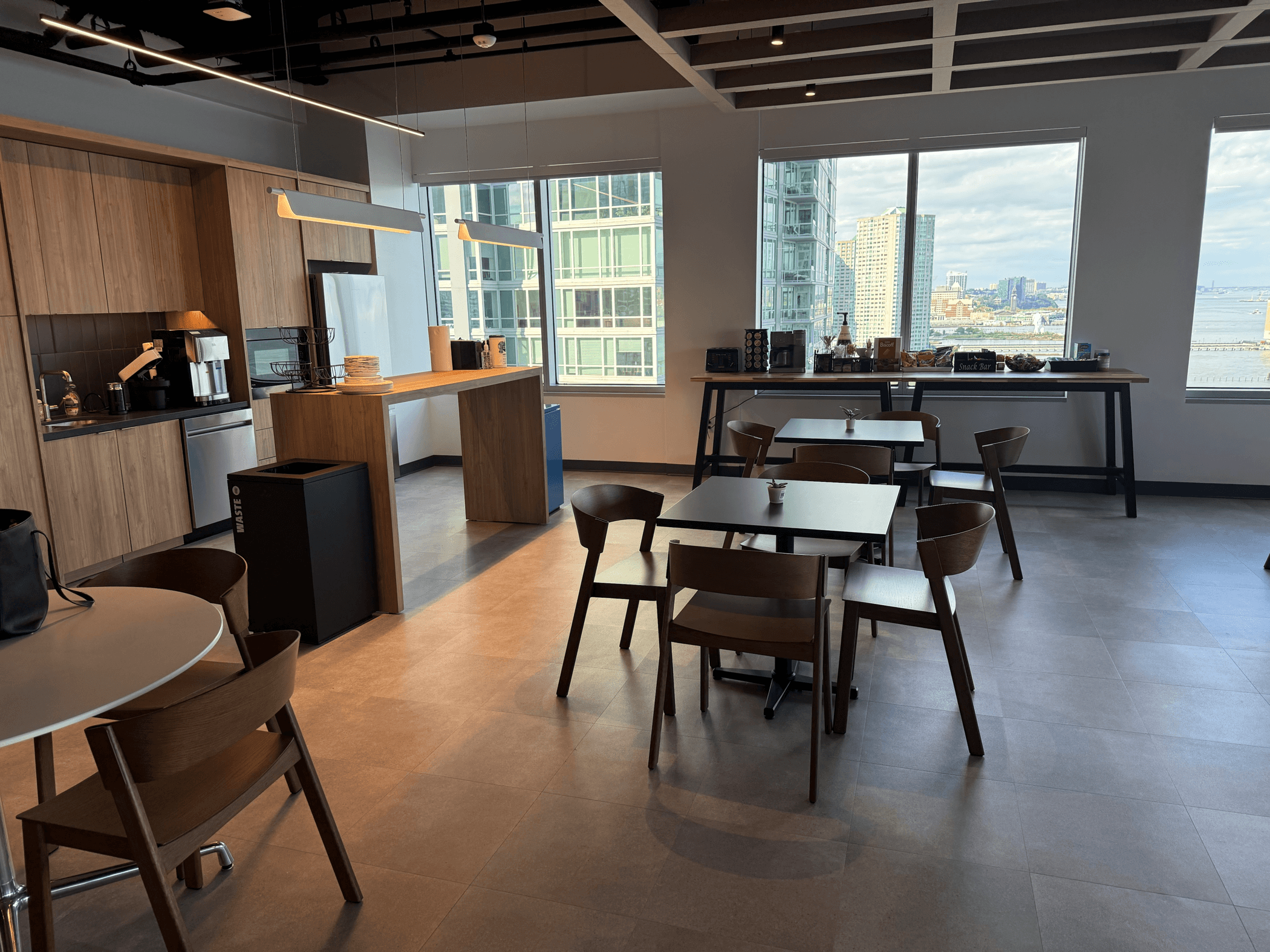 Modern office kitchen and café area with wood-finished cabinetry, coffee machines, and a refreshment station. Several dining tables and wooden chairs are arranged throughout the space, while large windows provide natural light and views of nearby high-rise buildings and a waterfront. Contemporary pendant lighting and clean, minimalist décor create a bright and welcoming environment for employees to relax, eat, and connect.